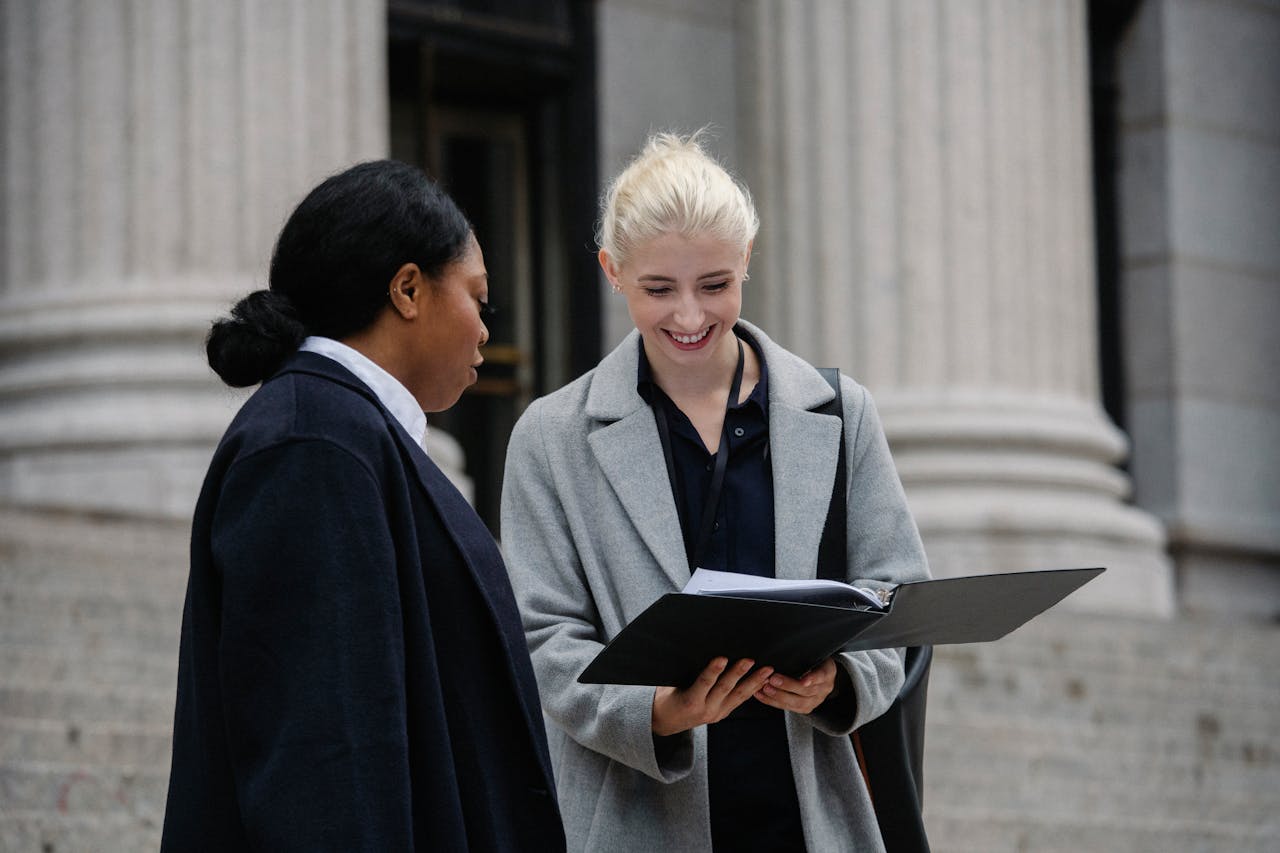 gallery-02 Smiling multiracial female coworkers wearing trendy formal outfits standing outside stone building and reading documents while discussing business together