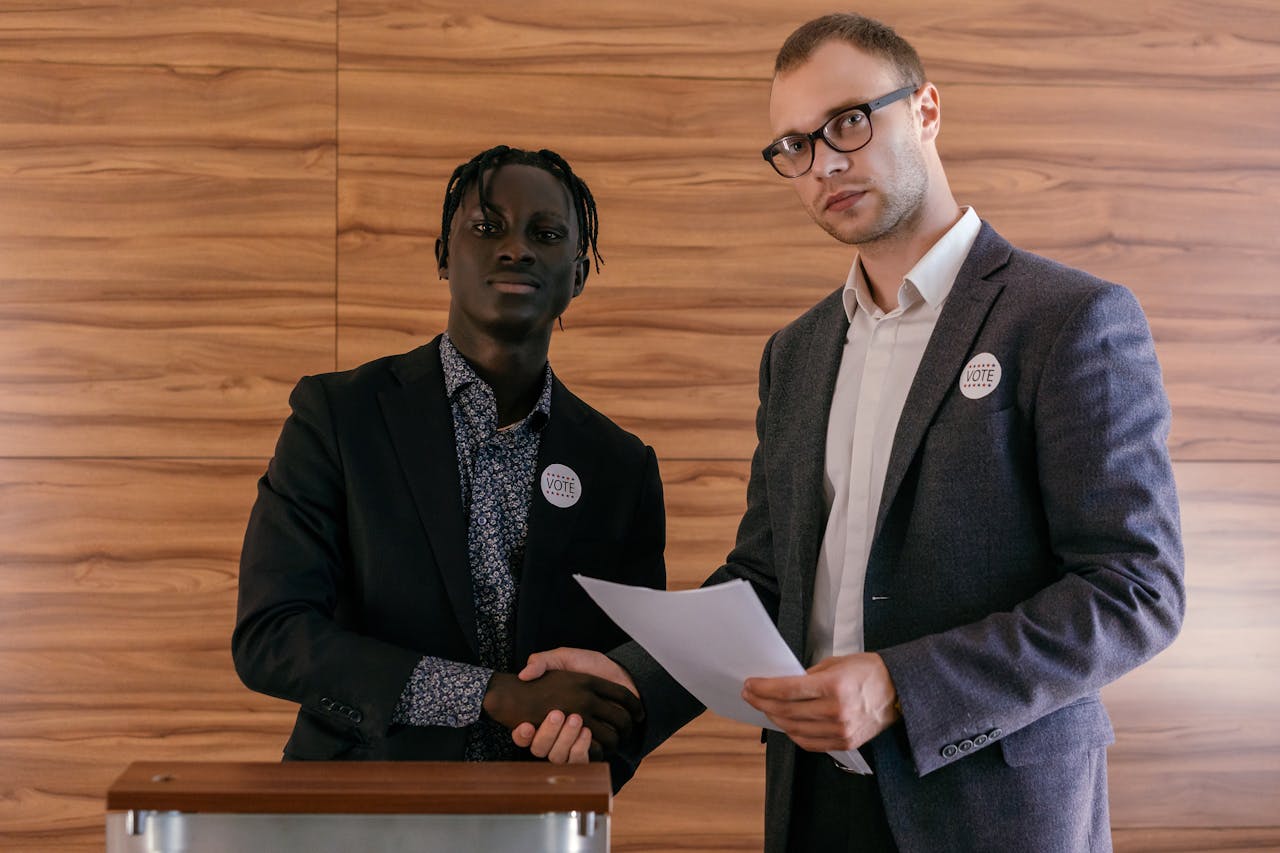 about-img-01 Two men in business attire shaking hands with agreement, promoting voting indoors.